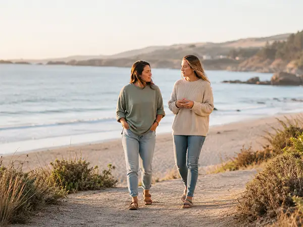 Two women walking and talking near a peaceful shoreline representing trauma therapy support and nervous system healing.