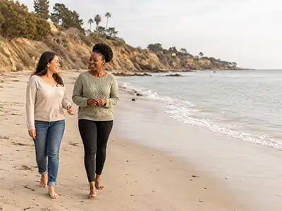 Hispanic woman and Black woman walking and talking along a peaceful beach during a walk and talk therapy session.